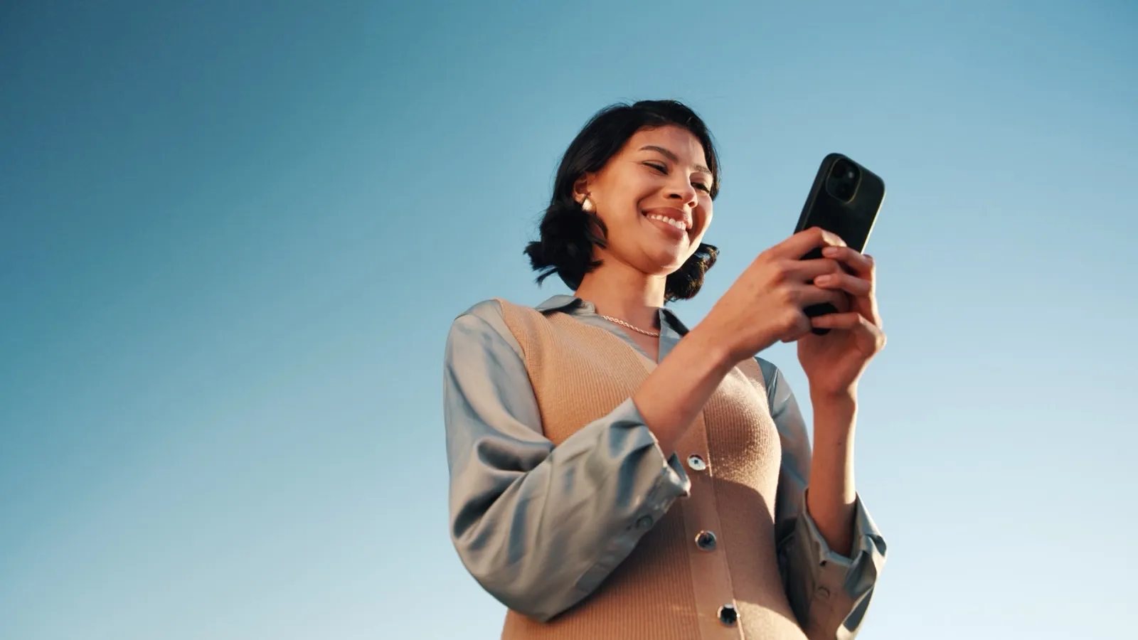 Mulher sorrindo ao olhar o celular, vista de baixo contra o céu azul.