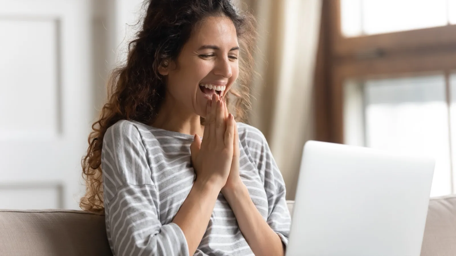 Mulher sorrindo de mãos juntas diante de um notebook, sentada no sofá.