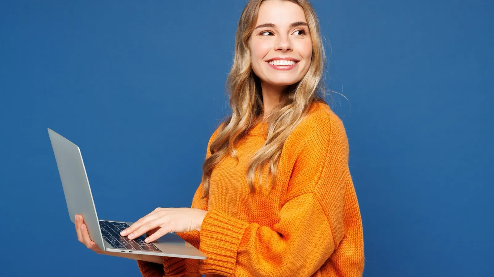 Mulher de suéter laranja sorrindo com um notebook aberto nas mãos, sobre fundo azul.