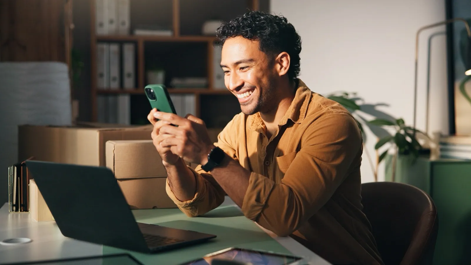 Homem sorrindo ao olhar o celular, sentado à mesa com notebook e caixas de encomenda ao lado.