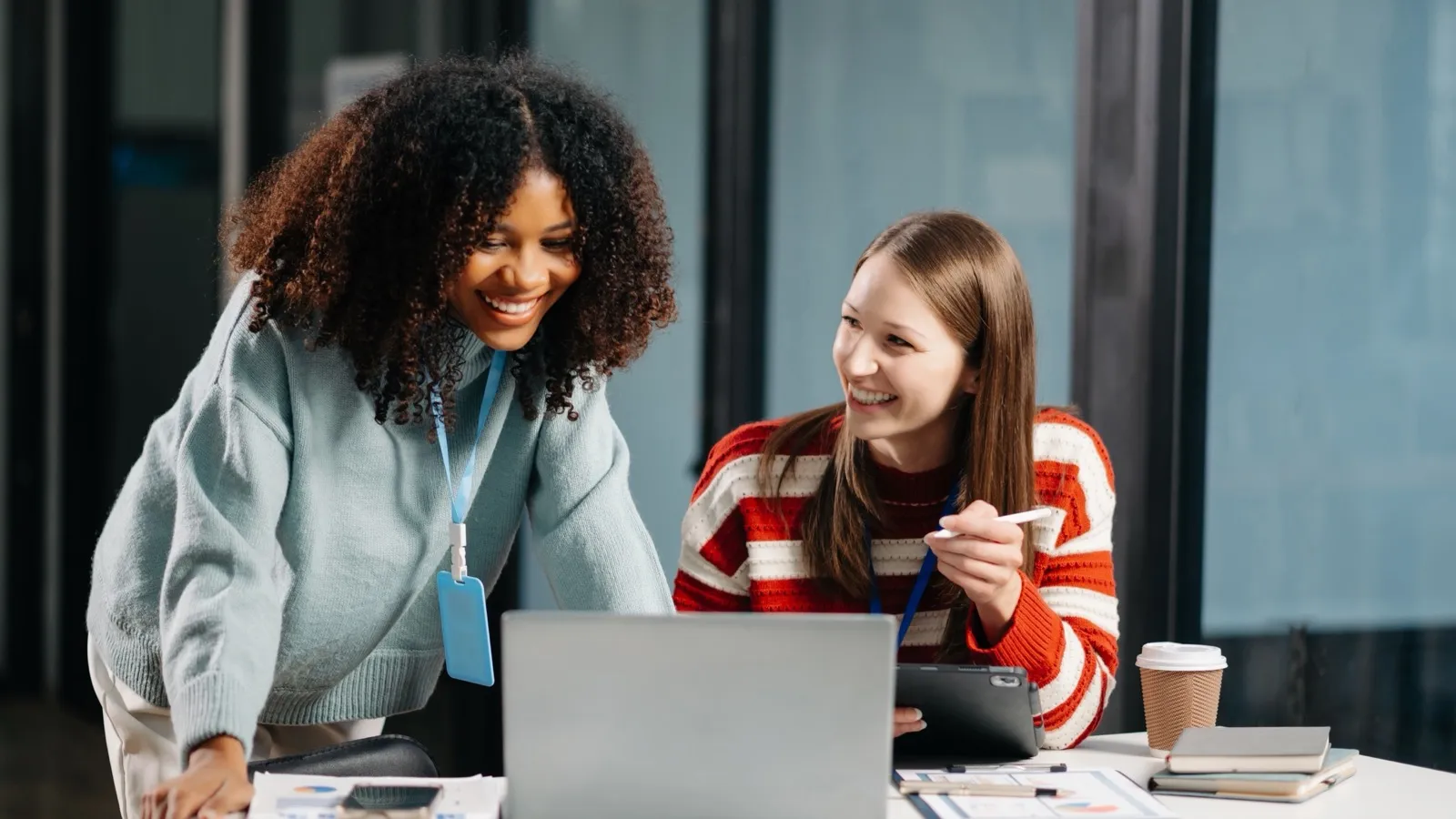 Duas colegas olhando juntas a tela de um notebook, sorrindo, numa mesa com relatórios impressos.