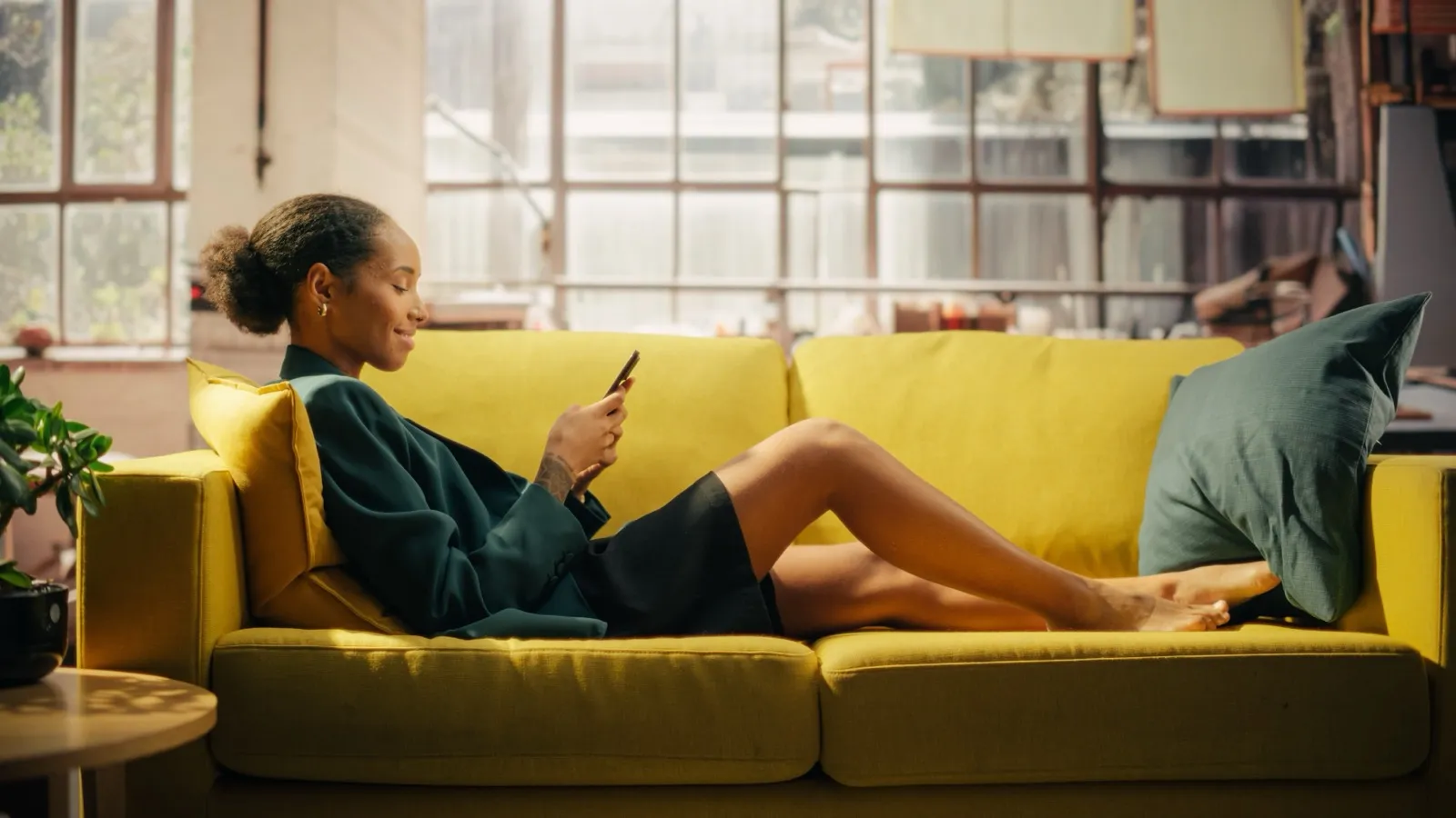 Mulher deitada num sofá amarelo olhando o celular, numa sala iluminada.