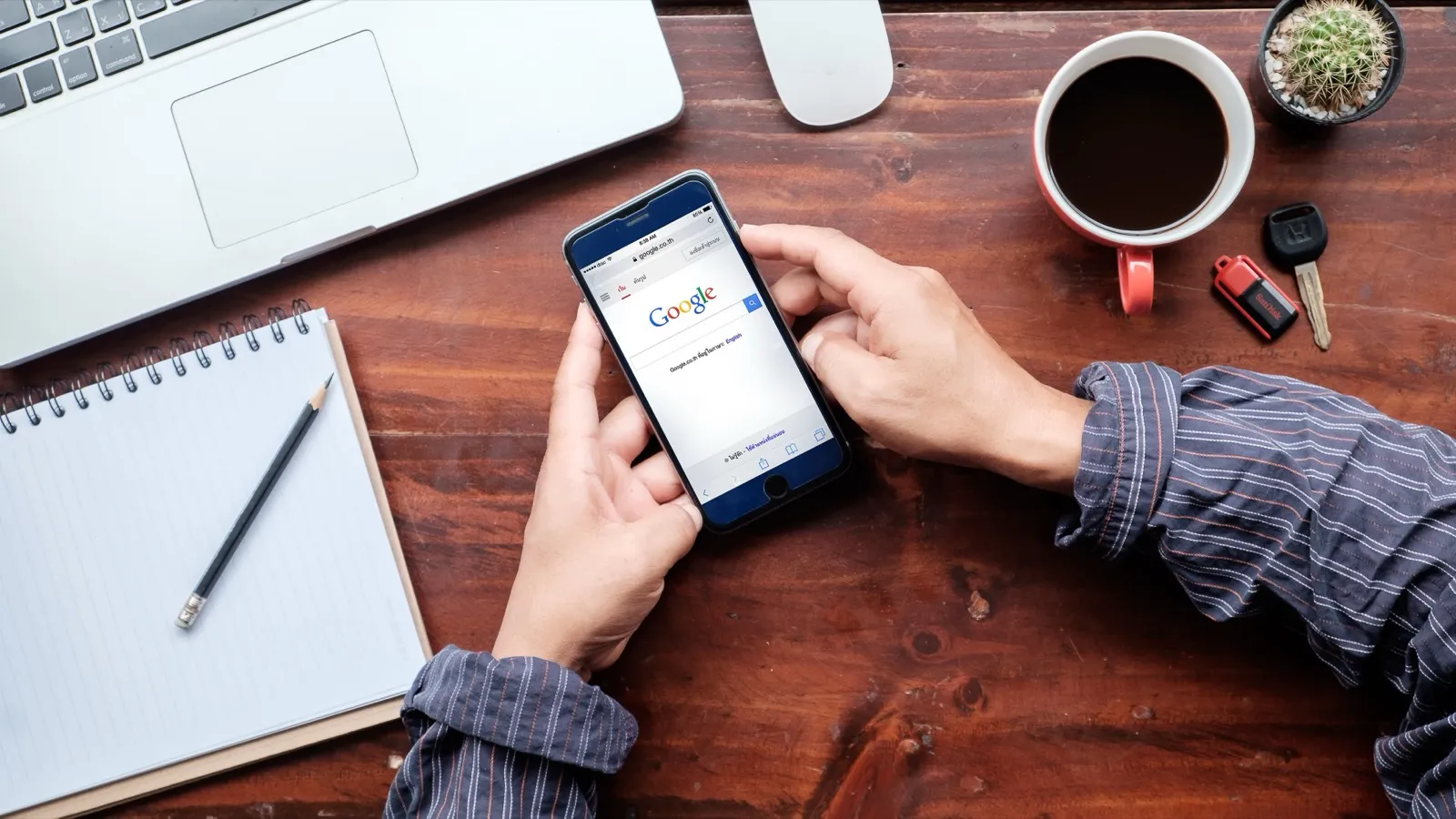 Pessoa segurando um celular com a busca do Google aberta, sobre uma mesa de madeira com notebook, caderno e xícara de café.